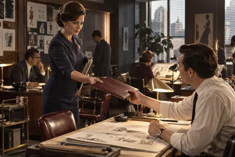 A woman passes a burgundy folder to a colleague seated at a drafting desk.
