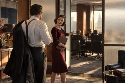 A woman gestures toward a meeting room as an arriving man removes his jacket.