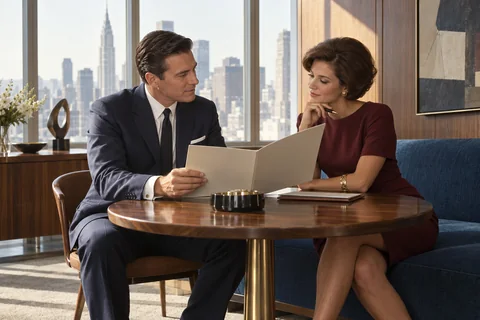 Two colleagues discuss a presentation folder at a round table beside city windows.