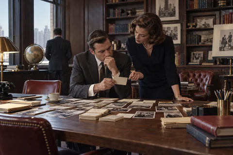 A man and woman study a small card among photographs and research papers spread across a table.
