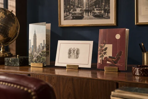 Three illustrated printed pieces stand on brass holders atop a walnut cabinet.