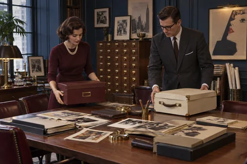 A woman and man set burgundy and cream archive boxes beside research materials on a table.