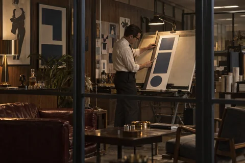 A designer trims a tall paper layout at an angled drawing board, seen through an office glass partition.