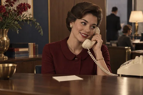 A smiling receptionist speaks on a cream rotary telephone behind a walnut counter.