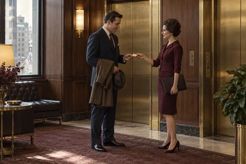 Two colleagues exchange a small card beside brass elevator doors.