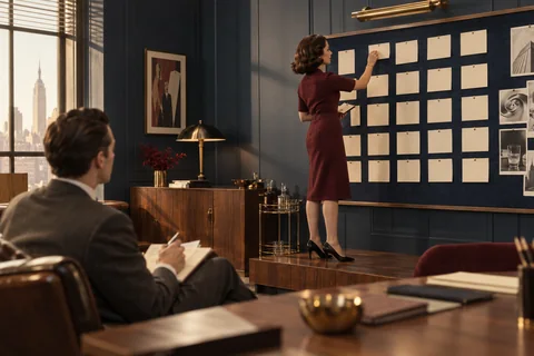 A woman pins a cream card to a planning board while a seated colleague takes notes.