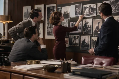 Four colleagues study black-and-white workplace photographs pinned to a large presentation board.
