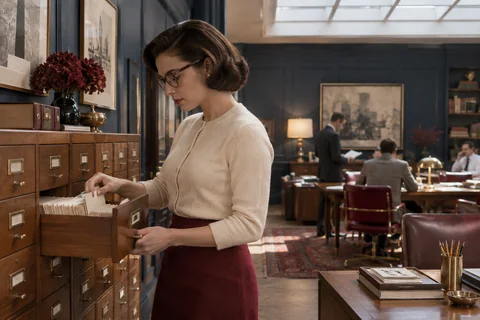 A woman in glasses selects an index card from an open walnut cabinet drawer.