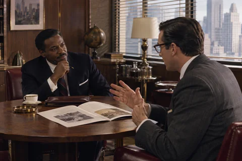 Two men discuss an open illustrated brochure at a round walnut table beside a bright window.